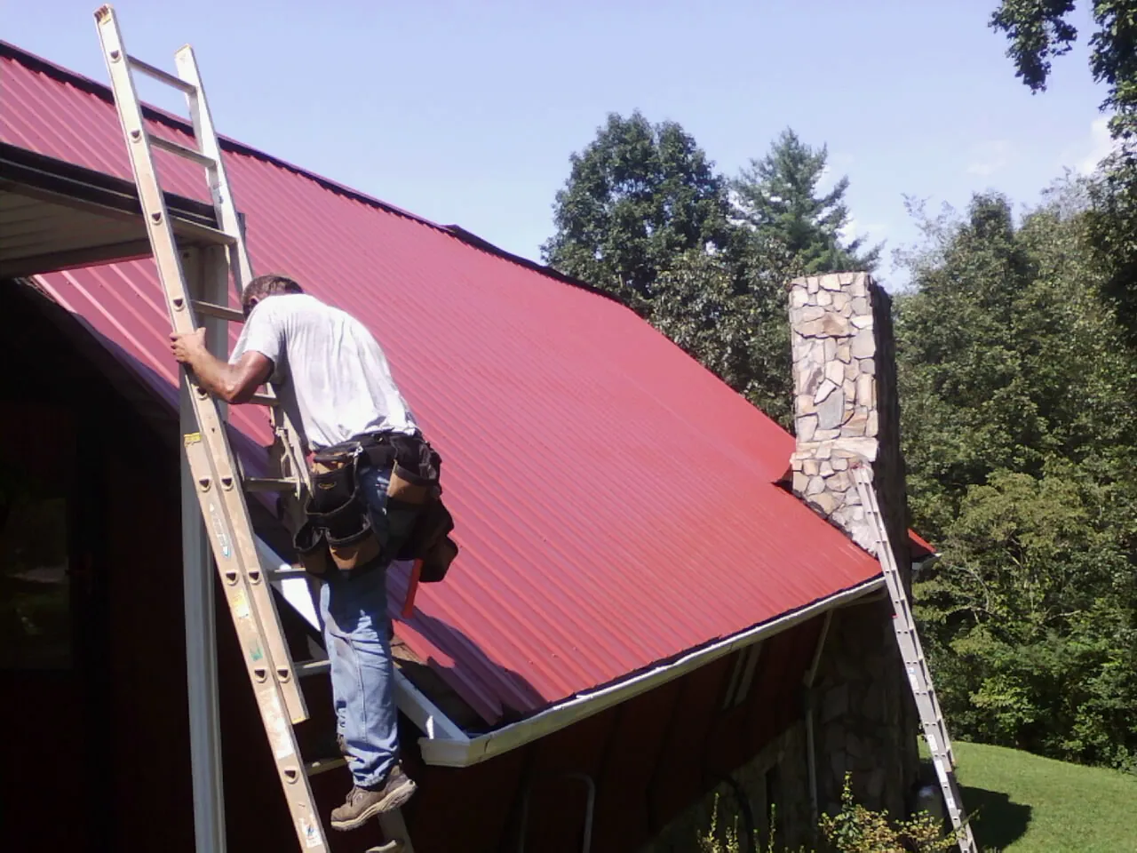 Roofer on a ladder beside a red metal roof and stone chimney — Soffit & Fascia Repair by Roofing Brothers in Albany